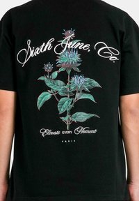 Svart t-shirt med en färgglad blommig grafik med gröna blad och blå blommor. Texten inkluderar "Sixth June, Co." och "Elevate Every Moment."