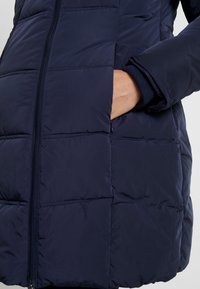 Navy blue puffer jacket with quilted texture, featuring a front zipper and side pockets. Soft fabric and ribbed cuffs enhance comfort.