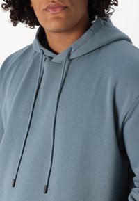 Hooded sweatshirt in a soft blue fabric, featuring a kangaroo pocket and drawstrings with black tips, smooth texture, and relaxed fit.