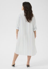 Robe chemise blanche avec des rayures verticales bleu clair, coupe décontractée, manches retroussées, atteignant mi-mollet, associée à des sandales à petit talon.