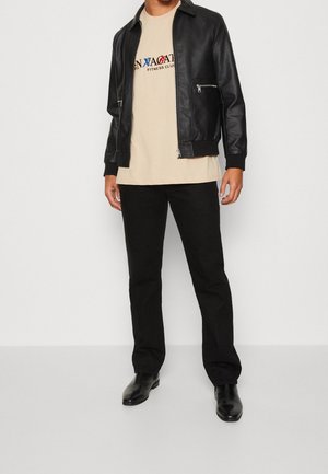 Czarna skórzana kurtka bomber z kieszeniami na zamek, beżowy t-shirt z tekstem tematycznym związanym z fitness, czarne spodnie i czarne botki za kostkę.