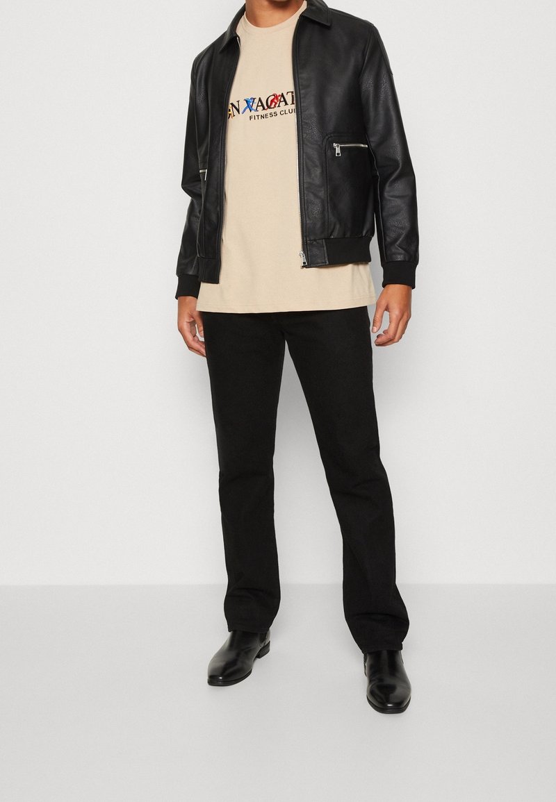 Veste bomber en cuir noir avec poches zippées, t-shirt beige avec un texte sur le fitness, pantalon noir et bottines noires.