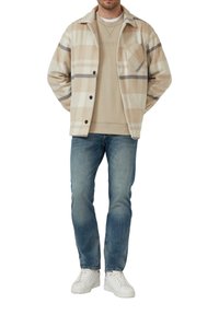 Béžová kockovaná bunda s tmavošedými pruhmi, zapínanie na gombíky a predná vrecko, nosená na béžovej mikine. Skombinovaná s modrými džínsy a bielymi teniskami.