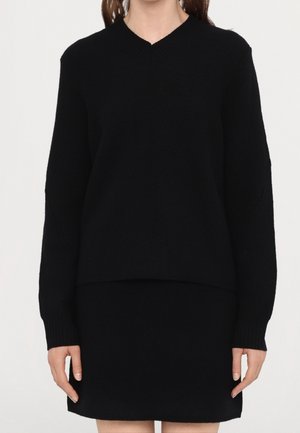 Femme portant un pull noir à manches longues avec col en V et une jupe noire assortie, debout devant un fond blanc uni.
