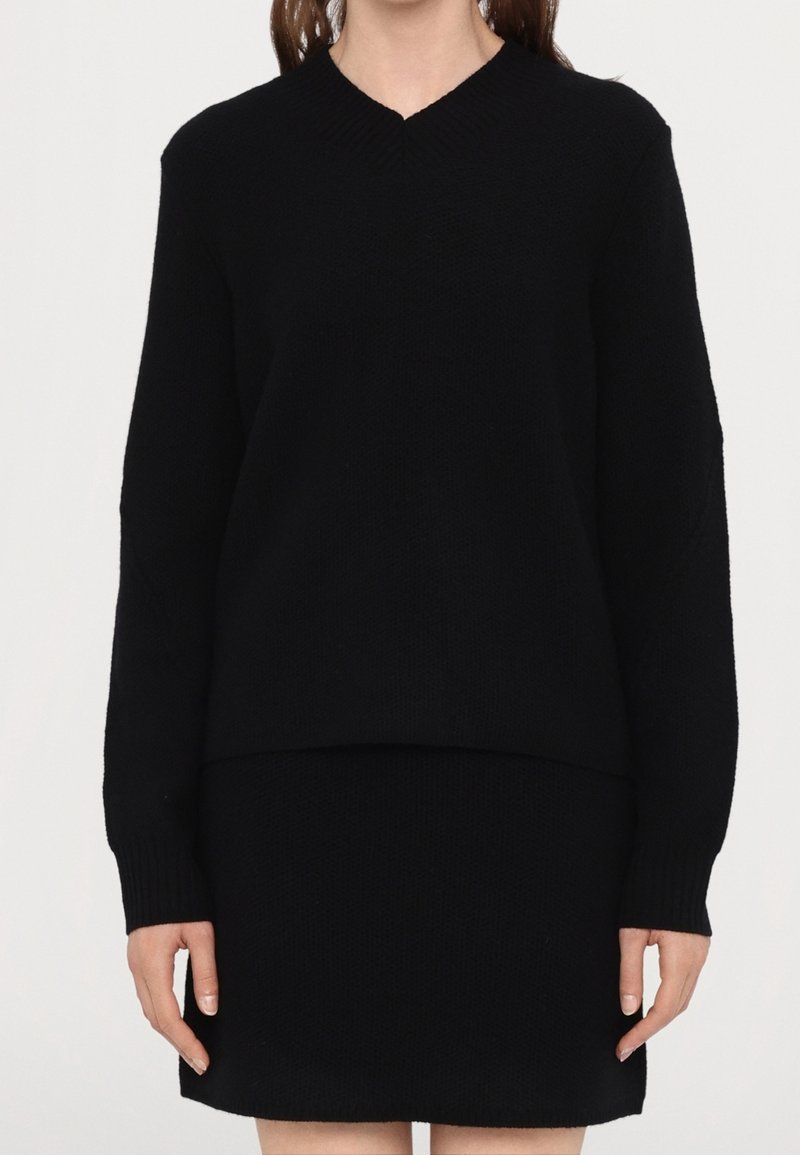 Femme portant un pull noir à manches longues avec col en V et une jupe noire assortie, debout devant un fond blanc uni.