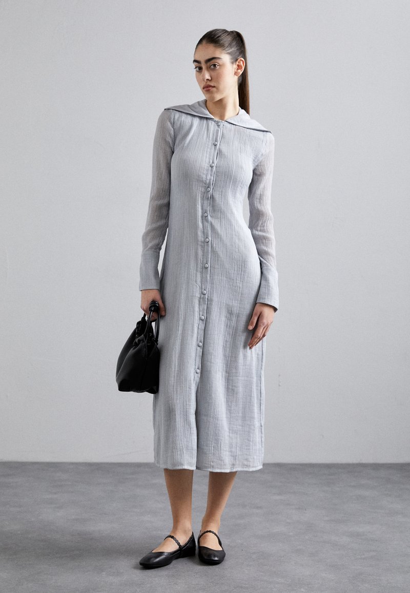Light gray button-up dress with a pleated texture, long sleeves, and a collar. Worn with black flats and a black handbag.