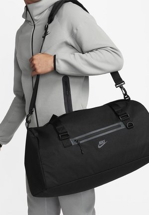 Sac de sport Nike noir avec bandeau réglable, forme cylindrique large, tissu texturé et un compartiment avant zippé.