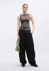 Haut gris sans manches, pantalon cargo noir surdimensionné et petit sac à main vert; le mannequin pose avec une main dans la poche, les matériaux texturés visibles.