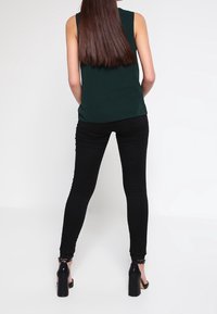Donna con lunghi capelli castani, indossa un top senza maniche verde scuro, pantaloni neri aderenti e sandali neri con tacco alto, in piedi su uno sfondo bianco.