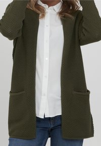 Olivově zelený pletený kardigan s texturovaným vzorem, otevřený vpředu, dlouhé rukávy a dvě přední kapsy, nošený přes bílé tričko a džíny.