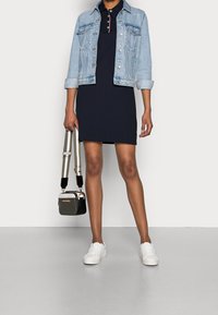 Mörkblå poloklänning med en ljus denimjacka, vita sneakers och en liten svart handväska med guldaccent. Slät tyg och avslappnad design.