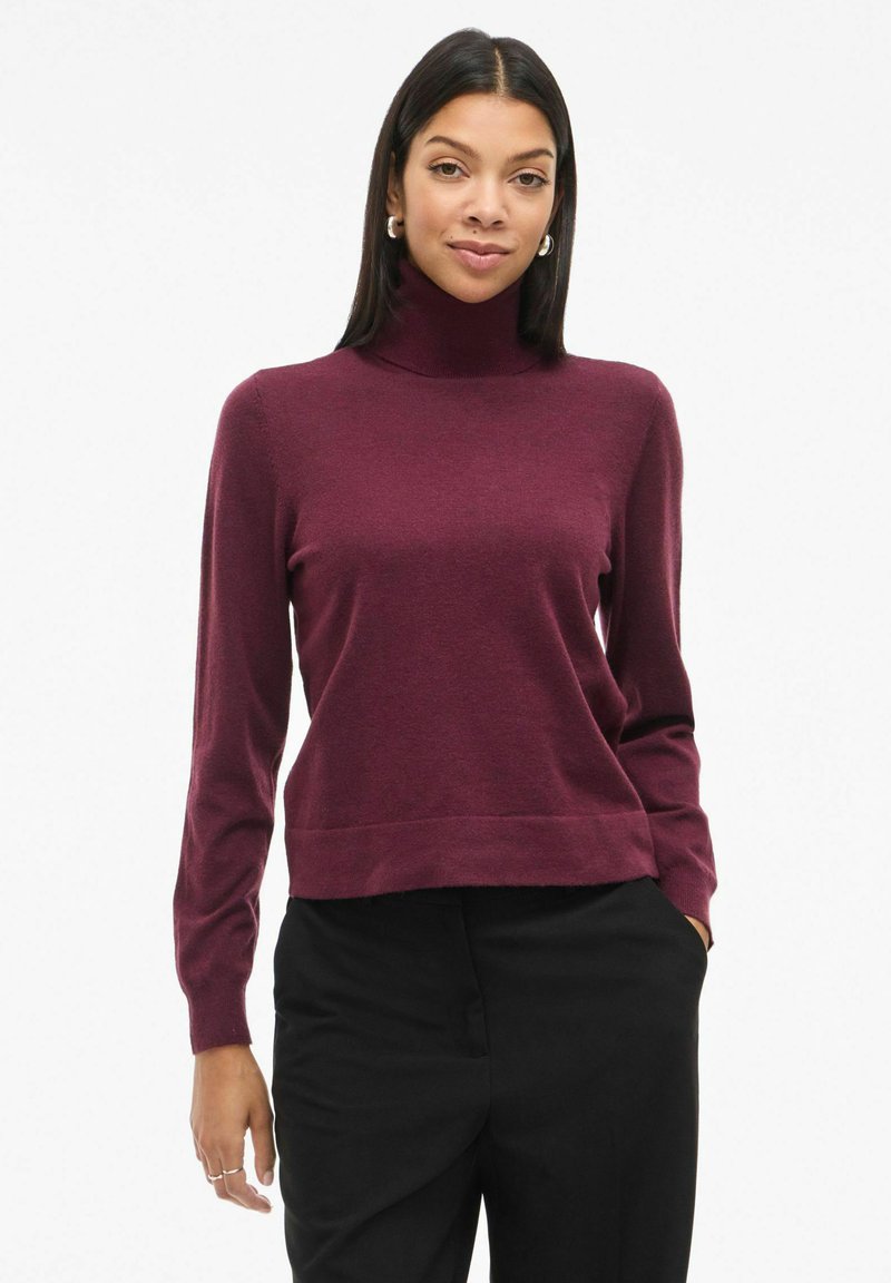 Burgundijas krāsu turtleneck džemperis ar garām piedurknēm, izgatavots no mīksta auduma, ar pieguļošu dizainu un īsu apakšmalu. Nēsāts ar melnām biksēm.