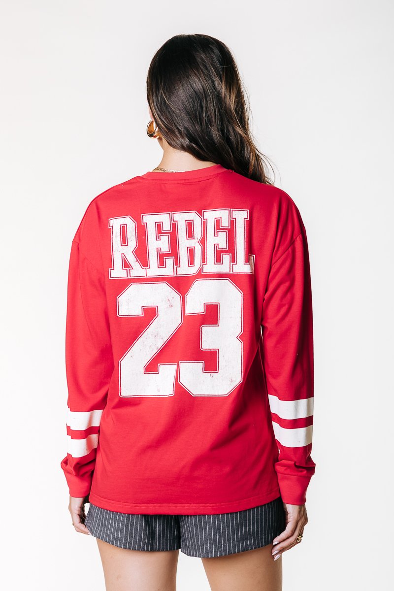 Červená košeľa s dlhým rukávom a bielym textom "REBEL" a grafikou "23" na zadnej strane. Obsahuje biele pruhované detaily na rukávoch. Vyrobená z jemného bavlneného materiálu.