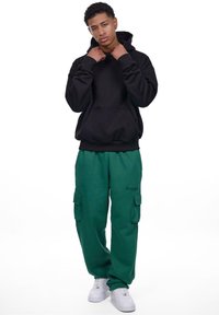 Svart hettegenser med kengurulomme, kombinert med grønne cargo-bukser med sidelommer og broderte tekster. Brukt med hvite joggesko.