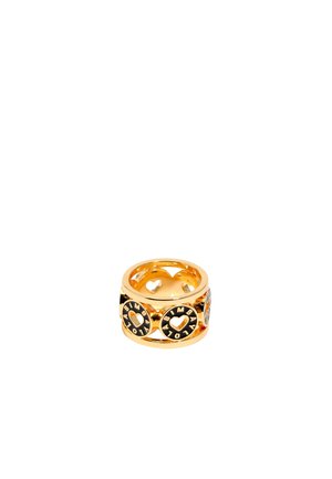 Anillo en tono dorado con corazones recortados y acentos circulares, que presenta detalles y inscripciones en negro. Superficie de metal lisa y pulida.