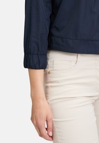 Veste légère bleu marine avec des manches froncées, associée à un pantalon crème comportant une poche à pièce et des rivets dorés.