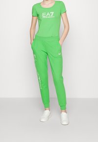 Zielony sportowy komplet z bawełny, składający się z dopasowanego topu z krótkim rękawem i zwężanych dresów z detalem z logo, zestawiony z białymi trampkami.