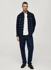 Camicia di flanella a quadri blu navy con bottoni bianchi, indossata sopra una t-shirt bianca a girocollo. Abbinata a jeans dritti blu scuro e scarpe nere.