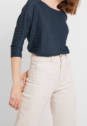 Kvinde iført navyblå tekstureret langærmet top og lyse beige bukser med høj talje og forlommer, stående mod en ensfarvet baggrund.