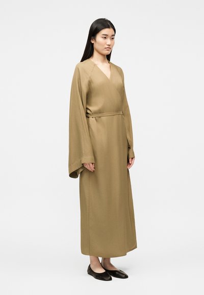 Femme debout portant une robe longue et ample de couleur beige avec de larges manches et une ceinture fine, associée à des chaussures plates noires.