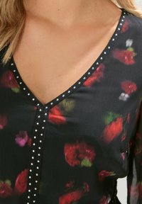 Blouse noire avec un motif floral multicolore. Design en V avec une ligne de clous en argent le long de l'encolure et un détail de bouton à l'avant.