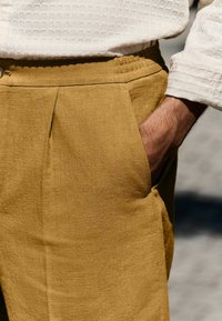 Beige linnedragt med en struktureret overflade, der har folder og sidelommer, parret med en hvid struktureret skjorte.