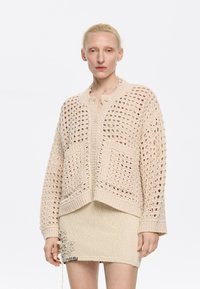 Cardigan all'uncinetto beige con vestibilità ampia, dotato di grandi tasche, chiusura a bottoni e polsini a costine. Indossato sopra una mini gonna beige strutturata.