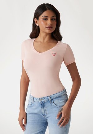 T-shirt rosa chiaro con scollatura a V, dalla forma aderente e maniche corte, con un piccolo logo triangolare rosso sul petto. Indossato con jeans azzurri chiari.
