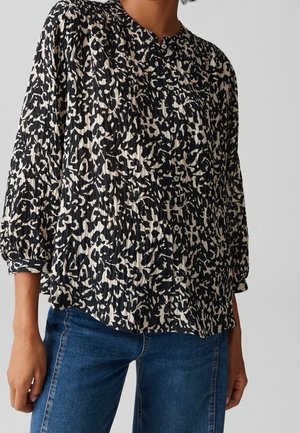 Femme portant un chemisier à boutons avec un motif abstrait noir et blanc, manches trois-quarts froncées et jean bleu.