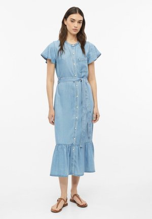 Frau steht und trägt ein hellblaues Jeans-Midikleid mit Knöpfen, kurzen Volantärmeln, einem Taillengürtel und braunen flachen Sandalen.