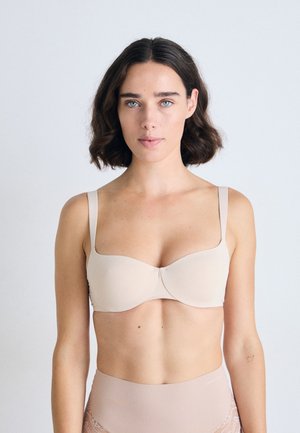 Kvinne med skulderlangt mørkt hår iført en beige BH og høyhåndet beige truse med blonder, står foran en enkel lys bakgrunn.