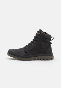Bota alta negra y resistente con suela texturizada, parte delantera con cordones, cuello acolchado, tirador trasero y logotipo circular de la marca en el costado.