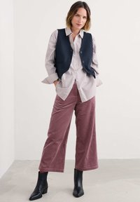 Námornícky vest over prúžkovanou košeľou na gombíky, v kombinácii s širokými, žebrovanými burgundskými nohavicami a čiernymi členkovými čižmami, na neutrálnom pozadí.