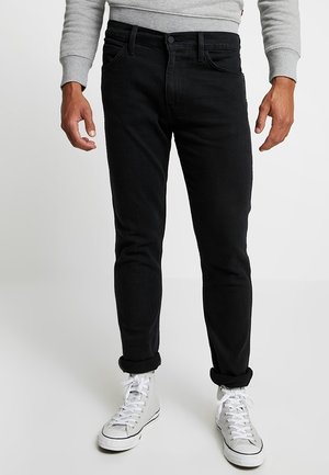 Man die zwarte slim-fit jeans met opgerolde pijpen draagt, een grijs shirt met lange mouwen en witte high-top sneakers, staat tegen een effen achtergrond.