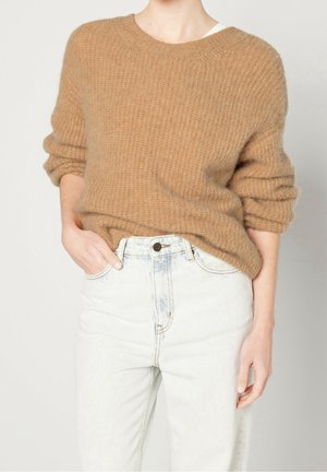 Osoba ubrana w luźny, teksturowany sweter w kolorze wielbłądziej wełny z podwiniętymi rękawami oraz wysokiej talii jasne dżinsy z przetarciami, z ręką w kieszeni.