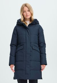 Marineblå pufferjakke med avtagbar hette, glidelås foran, to sidelommer og quiltet tekstur. Glatt stoff og knelengde design.