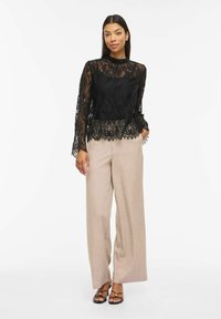 Femme portant un chemisier noir à manches longues en dentelle, un pantalon large beige et des sandales à talons noires à brides, debout avec la main dans la poche.