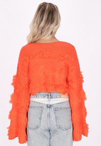 Helle orange, kurz geschnittene Pullover mit fransenverzierten Ärmeln und Körper, kombiniert mit hochtaillierten Jeansshorts mit einem Etikett auf der Rückseite.