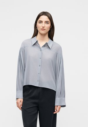 Femme portant un chemisier gris clair boutonné avec des poignets larges et un pantalon noir, debout devant un fond blanc uni.