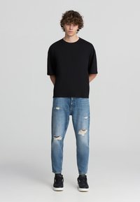 T-shirt nero a maniche corte abbinato a jeans chiari strappati. I jeans presentano numerosi strappi e sono indossati con sneakers nere.