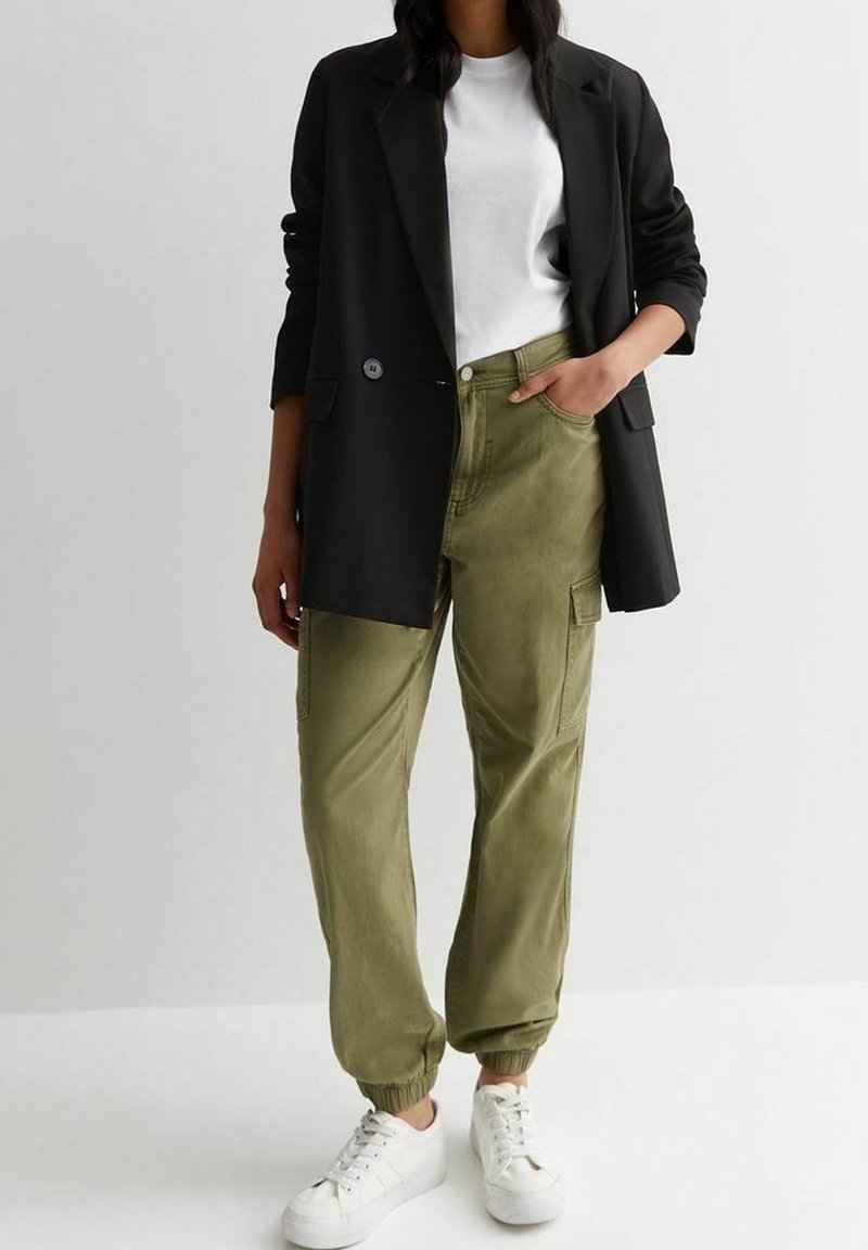 Sort blazer over en hvid t-shirt, matchet med olivengrønne cargo-bukser med elastiske manchetter. Hvide sneakers fuldender outfittet.