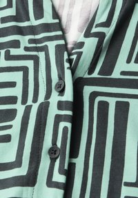 Knopfjacke aus mintgrünem Stoff mit schwarzen geometrischen Mustern. Auffällige schwarze Knopfdetails und strukturierte Strickqualität.