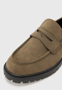 Loafer maro din suede cu vârf rotund, detaliu decorativ sub formă de curea și o talpă groasă, texturată din cauciuc negru pentru durabilitate.