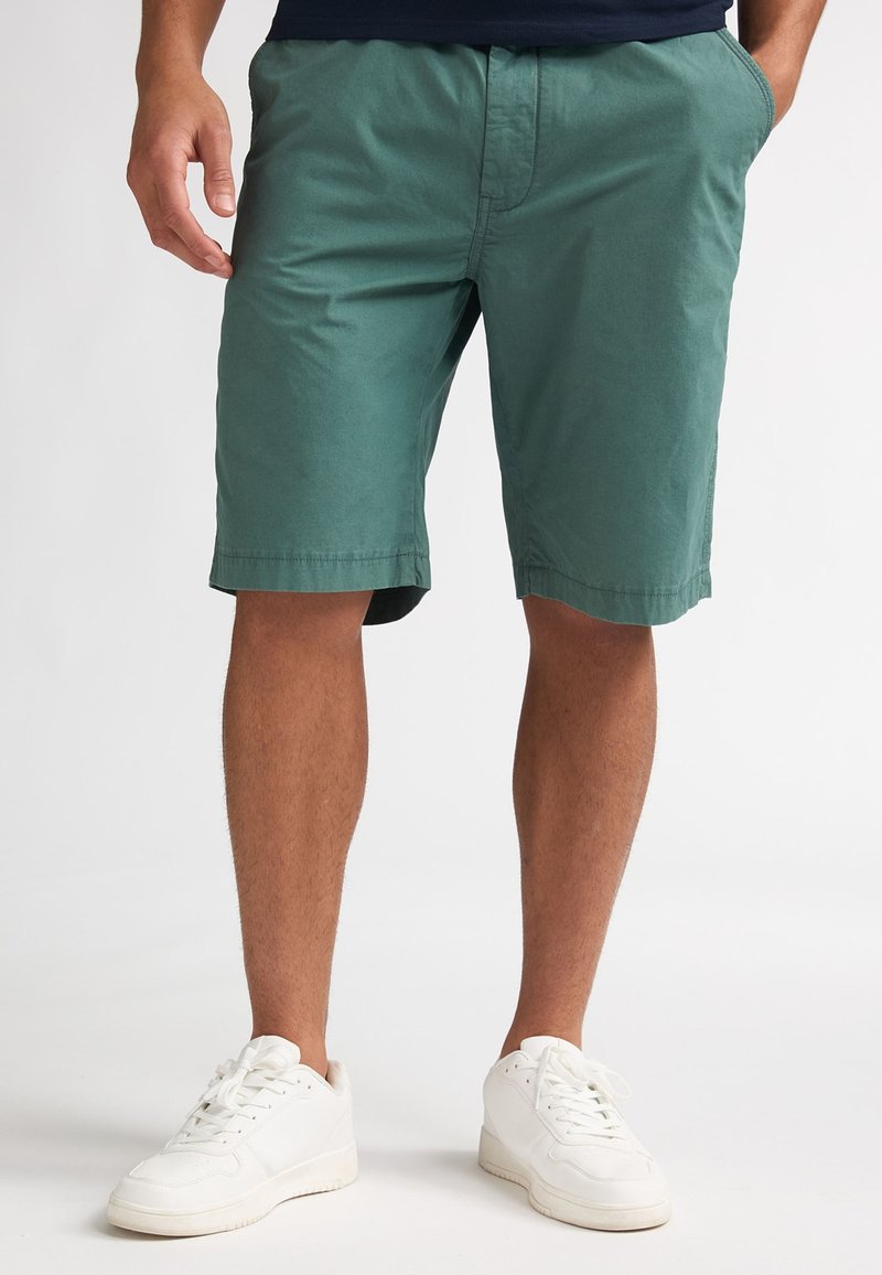 Homme portant un short vert jusqu'aux genoux et des baskets blanches, debout les mains dans les poches devant un fond uni.