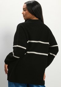 Pull noir à côtes avec des rayures horizontales blanches, présentant un col haut, des manches longues et une coupe décontractée. Matériau texturé.