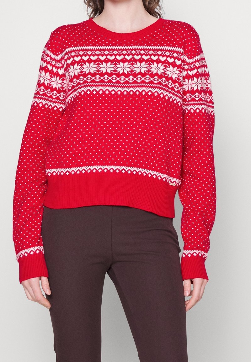 Pull en tricot rouge avec des motifs Fair Isle blancs. Il présente un col rond et des poignets côtelés. Porté sur un pantalon marron.