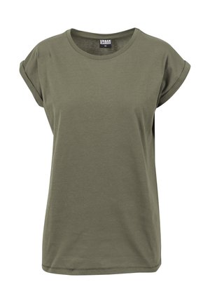 Kobieta ubrana w oliwkowozielony t-shirt z krótkim rękawem i jasnobeżowe spodnie, stojąca na tle gładkiego jasnoszarego tła.