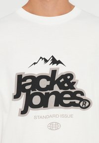 Camisa de algodón blanca con un gran logo negro que incluye "jack&jones", un gráfico de montañas, y el texto "STANDARD ISSUE" en gris debajo.