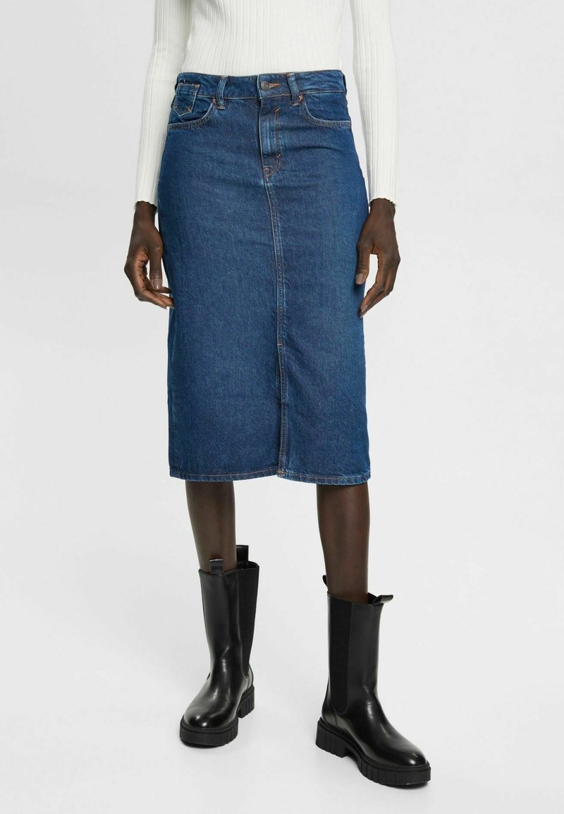 Femme portant une jupe midi en denim bleu, un haut blanc à manches longues côtelé, et des bottes noires épaisses mi-mollet, debout sur fond blanc.