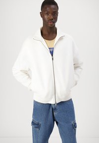 Hvit glidelåsgenser med høy krage og ribbestrikkede mansjetter, kombinert med blå denim cargobukser med sidelommer.
